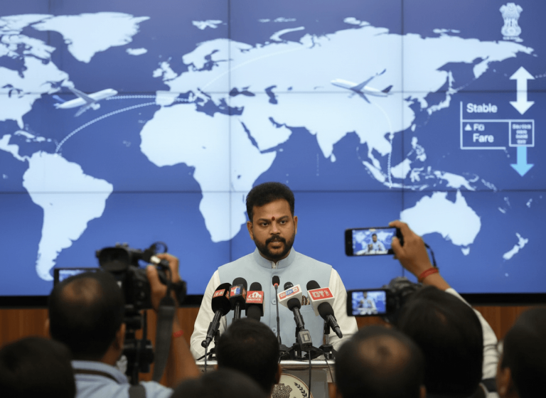 A man in a blue vest speaks at a podium with microphones, a world map with flight paths on screens behind him.