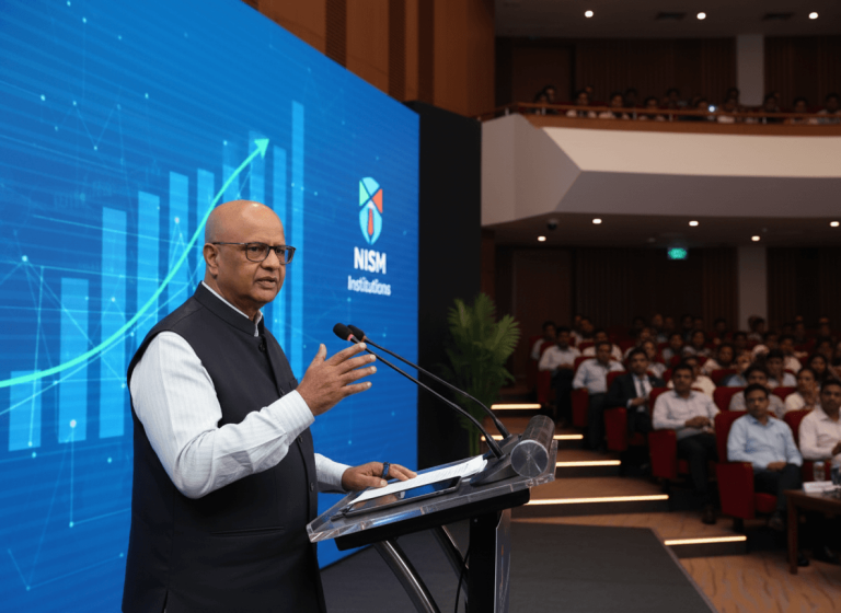 A bald man in glasses and a dark waistcoat speaking at a podium in front of a large screen with a bar graph.