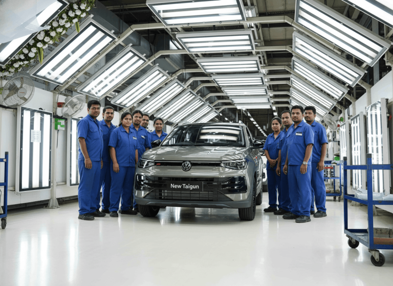 A group of factory workers in blue uniforms stand proudly around a new gray Volkswagen Taigun SUV on the assembly line.