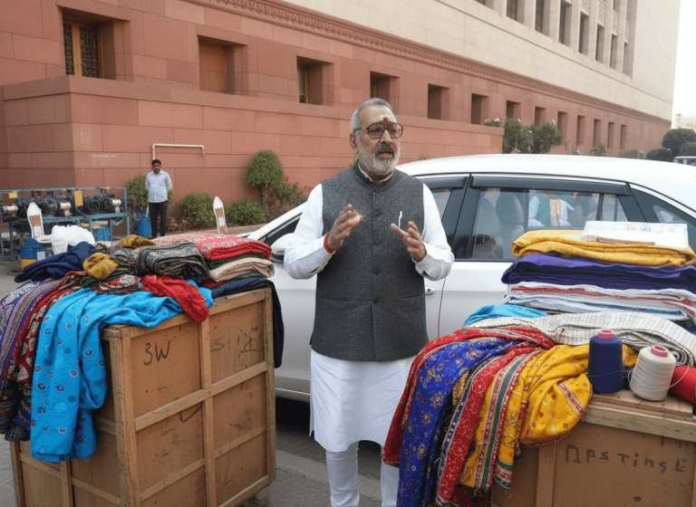 A man in traditional Indian attire gestures while standing near stacks of colorful fabrics and yarn outdoors.