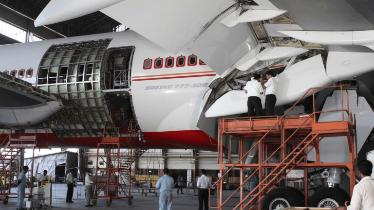 Wide shot of an Air India Boeing 777 in a hangar with technicians on platforms, working on the fuselage and wing.