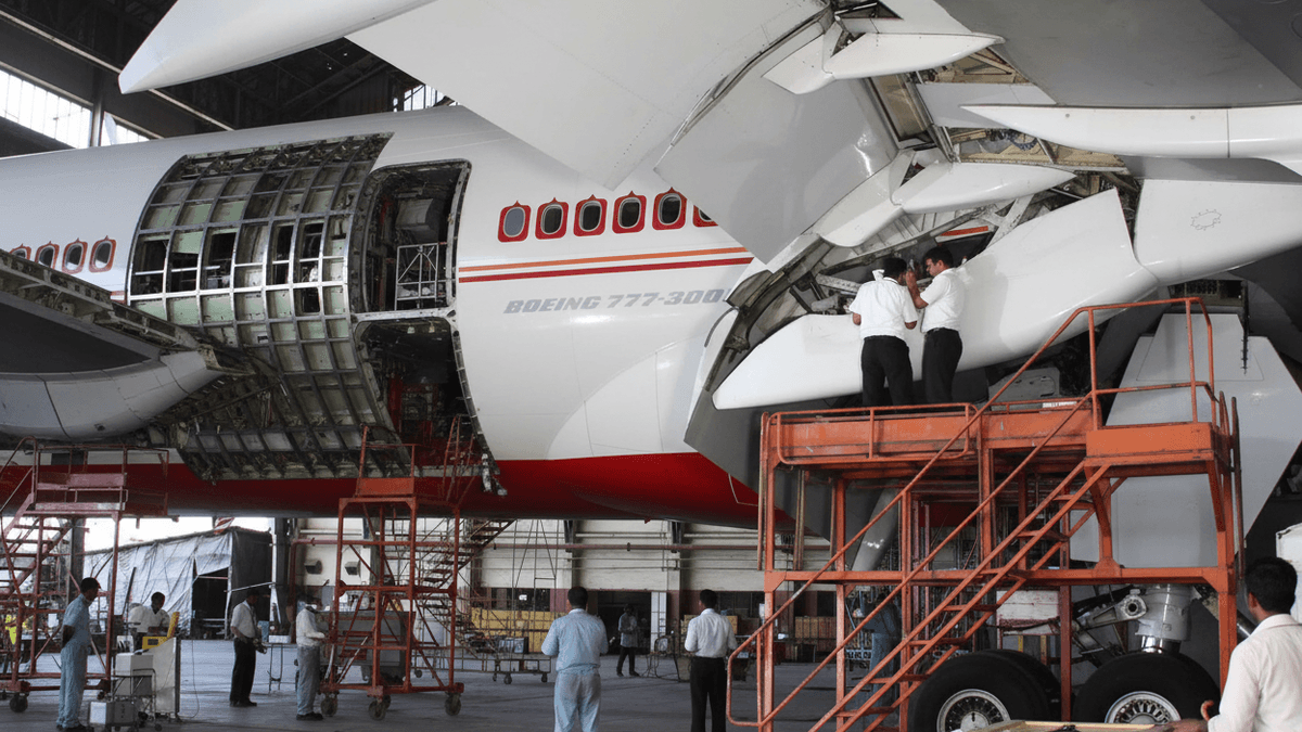 Wide shot of an Air India Boeing 777 in a hangar with technicians on platforms, working on the fuselage and wing.