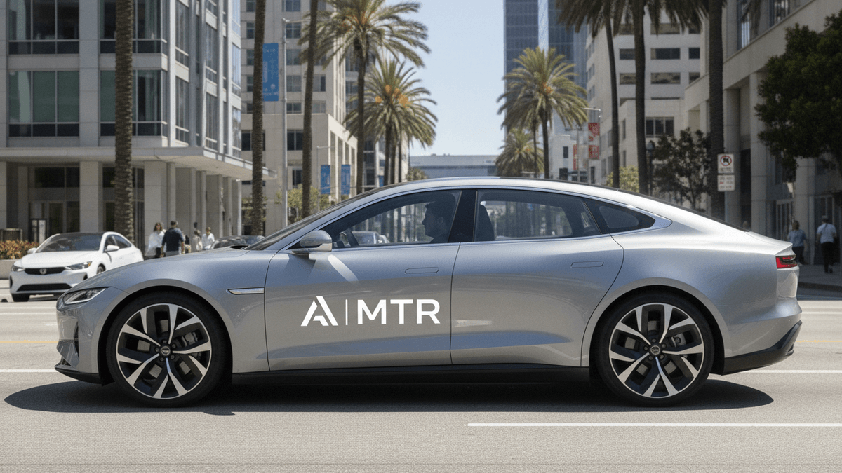 A silver modern sedan with a minimalist "A|MTR" logo on its side, driving on a sunny street in a California city.
